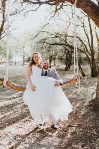 Hollow Hill Event Center Wedding and Event Venue, Weatherford, Texas. Bridge and groom sitting on swing made of a tree log. Trees all around