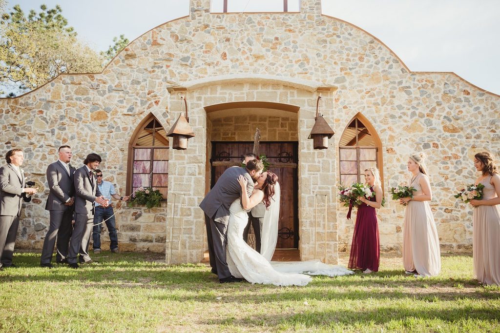 Hollow Hill Event Center Wedding and Event Venue, Weatherford, Texas. Bride and groom kissing in front of stone alamo style building. Wedding party around them.