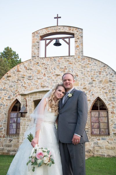 Hollow Hill Event Center Wedding and Event Venue, Weatherford, Texas. Bride and groom standing in front of stone alamo type building. Bride has head on grooms shoulder. Bouquet down at side.
