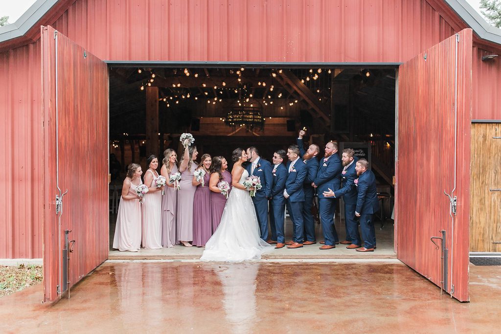 Hollow Hill Event Center Wedding and Event Venue, Weatherford, Texas. Bride, groom and wedding party standing just inside open double doors of red barn.