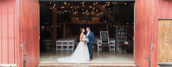 Hollow Hill Event Center Wedding and Event Venue, Weatherford, Texas. Bride and groom standing inside doorway of red barn building with doors open. Behind them are white chairs lined up