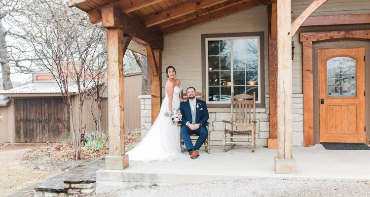 Hollow Hill Event Center Wedding and Event Venue, Weatherford, Texas. Bride standing beside groom sitting on porch of tan house.