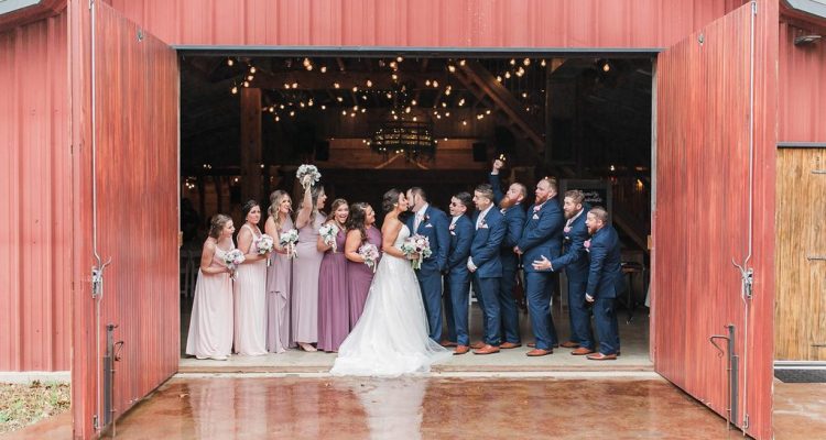 Hollow Hill Event Center Wedding and Event Venue, Weatherford, Texas. Bride, groom and wedding party standing just inside open double doors of red barn.